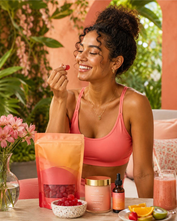 A woman in a coral pink sports bra sits at a table with skincare and wellness products, including a stand-up pouch of red berries, a jar, a dropper bottle, and a glass of pink drink, in a warm, tropical indoor setting.