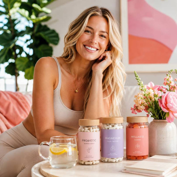 A smiling woman in a beige ribbed tank top and leggings sits indoors beside a round table holding three supplement jars labeled Probiotic, Magnesium, and Wellness, with a glass of lemon tea and flowers nearby.